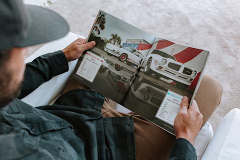 A person reads a Drivers Club magazine featuring two classic white cars with specifications and prices shown on the pages.