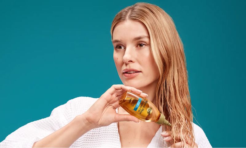 Woman applying Miracle Shine Argan oil from an amber bottle to the ends of her damp hair against a teal background.