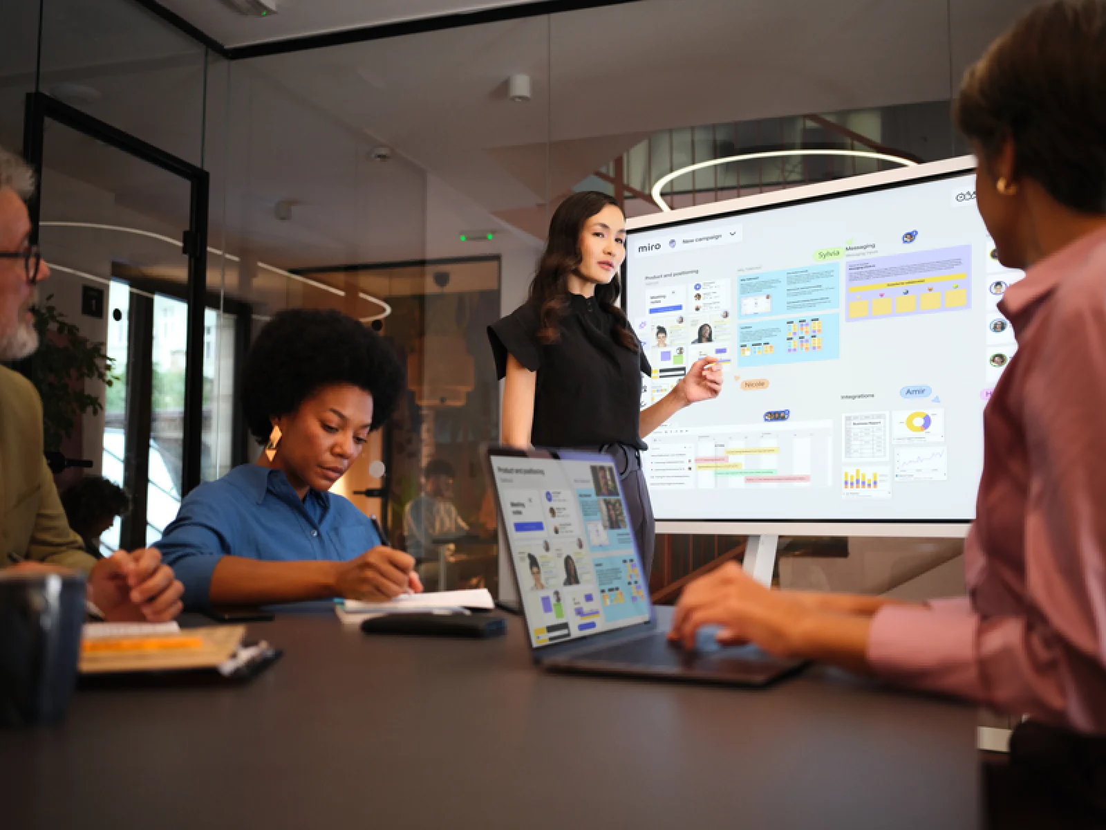 A woman presents a Miro board titled 'New campaign' to colleagues in a modern conference room, with the board displayed on a large screen showing product positioning content, user personas, charts, a Gantt-style timeline, and integrations panels, while a laptop on the table mirrors the same board.