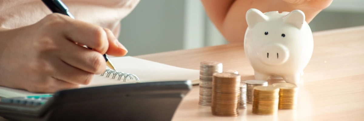 A person writing in a notepad, with some stacked coins and a piggy bank sitting nearby.