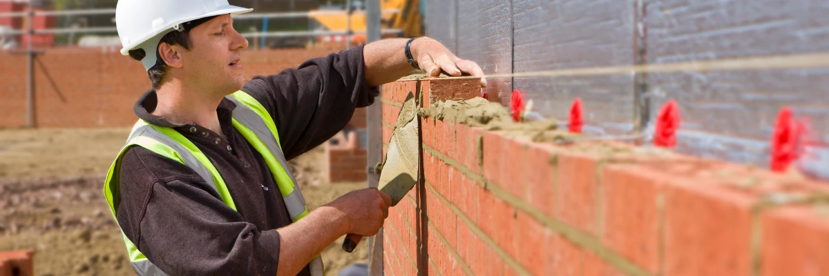 A bricklayer scraping mortar off a partly constructed brick wall.