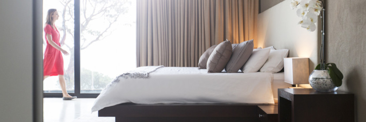 A woman walking through her bedroom, with a made bed furnished with pillows.