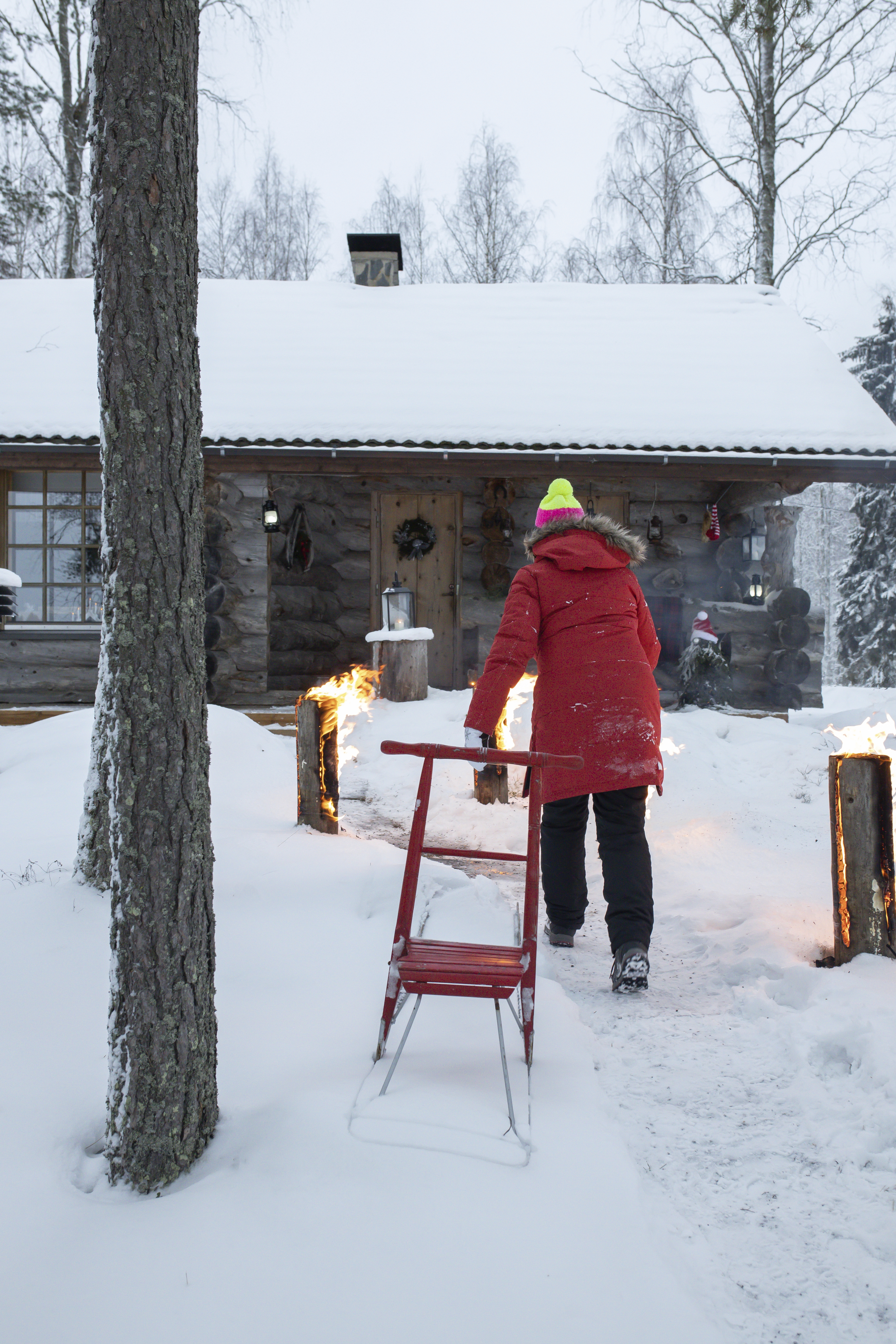 Nainen kävelee kohti kelomökkiä lumisella pihalla, jossa on punainen potkukelkka ja jätkänkynttilöitä.
