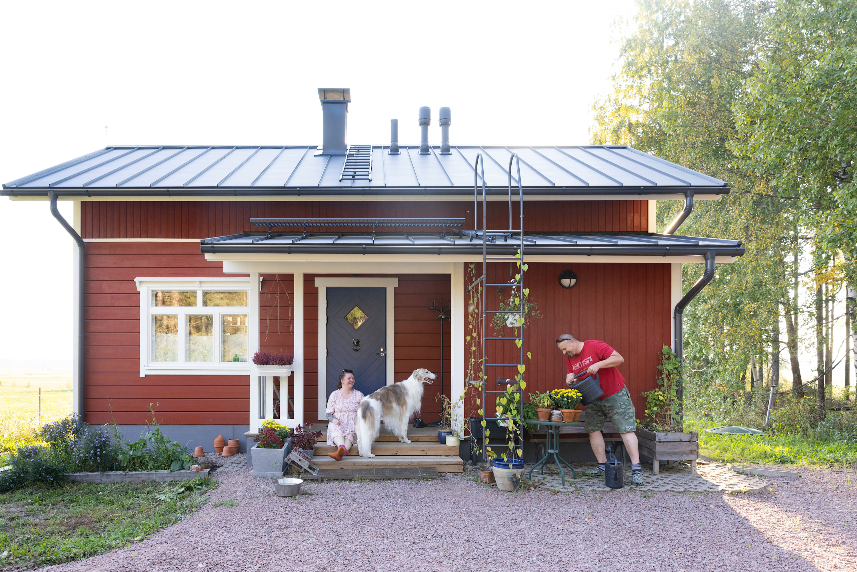 Tiia Inkinen punaisen talonsa terassilla koiran kanssa, puoliso kastelee kukkia.