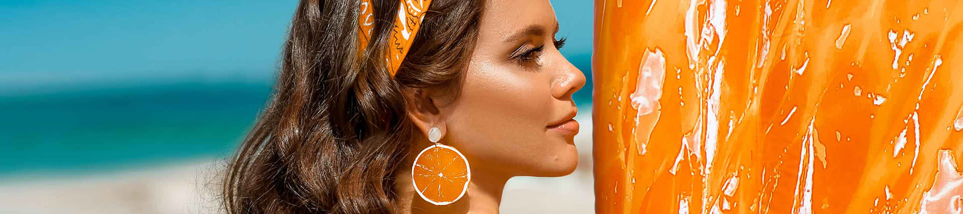 Back of brunette woman with orange bandana and a giant orange inflatable float looks to the right while standing on the beach