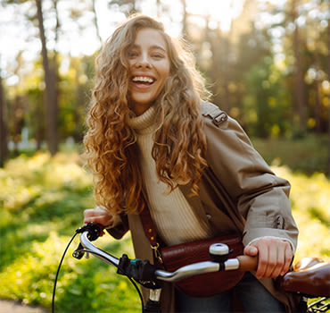 Happy pensive young woman in stylish clothes riding a bike in a sunny park, outdoors, looking away.