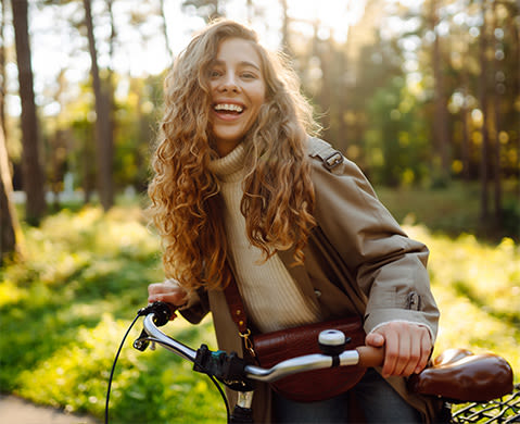 Happy pensive young woman in stylish clothes riding a bike in a sunny park, outdoors, looking away.