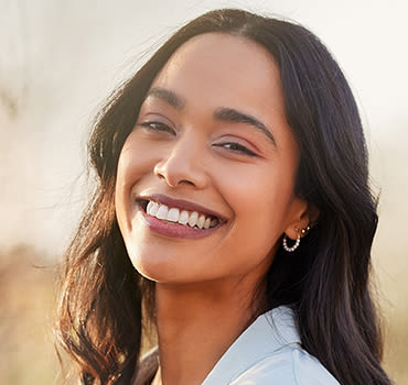 Woman with long dark brown hair smiling in front of an unfocused brown and green background