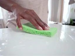 Hand wiping a white countertop with a green sponge