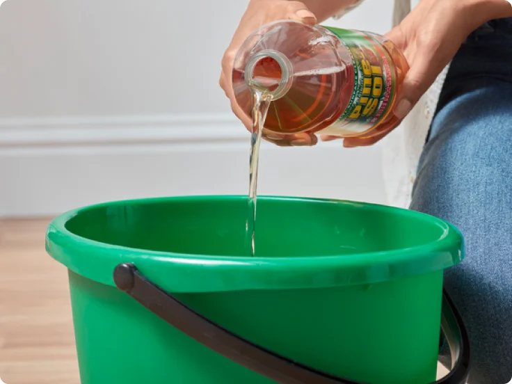 Pouring Pine-Sol Original Pine cleaner into a green plastic bucket.
