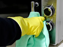 Yellow-gloved hand wiping the front of a microwave with a light green cloth