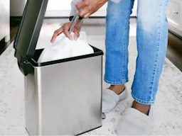 Persona con jeans y pantuflas pone bolsa blanca en bote de basura abierto de acero inoxidable en cocina.