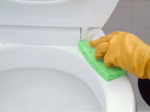 Mano enguantada utilizando una esponja verde para limpiar el borde de un inodoro.