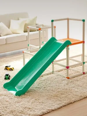 Kids' indoor green plastic slide on shag rug in living room, white sofa and toy cars in background.
