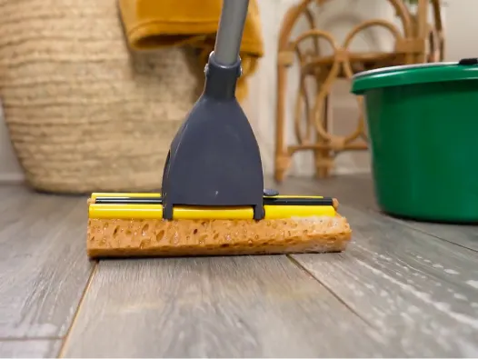 Primer plano de un cabezal de trapeador amarillo trapeando un piso laminado con un balde y una canasta verdes en el fondo