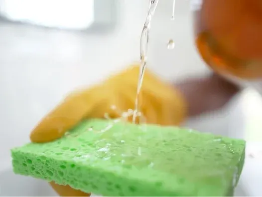 Mano enguantada de color amarillo que vierte un líquido transparente sobre una esponja verde