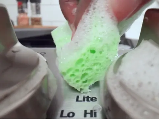 Close-up shot of sudsy green sponge cleaning stovetop controls.