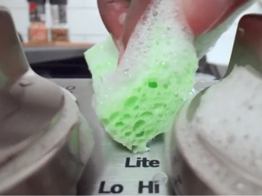 Close-up shot of sudsy green sponge cleaning stovetop controls.