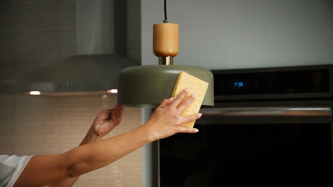 Hand cleaning a green hanging lamp in a kitchen with a yellow sponge