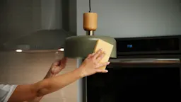 Hand cleaning a green hanging lamp in a kitchen with a yellow sponge