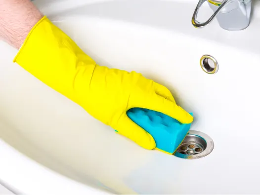Gloved hand using turquoise sponge to clean bathroom pedestal sink drain top.