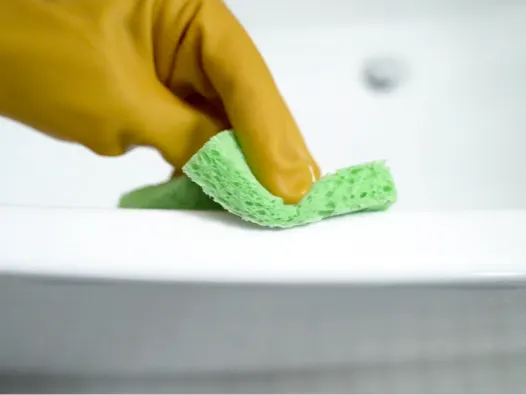 Mano enguantada usando una esponja verde para limpiar el borde de la bañera.