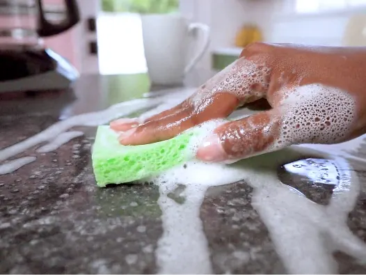 Mano y esponja verde limpiando una encimera de granito con burbujas de espuma