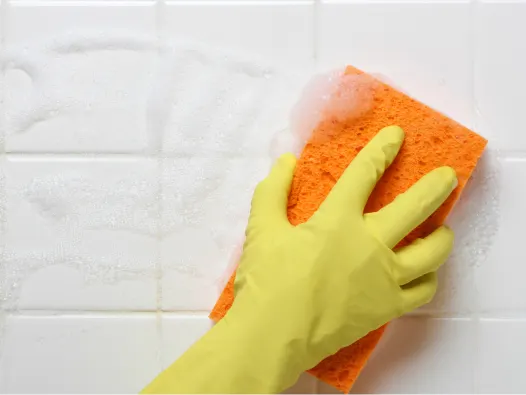 Mano enguantada de goma limpiando la pared de azulejos blancos de la ducha con una esponja naranja.