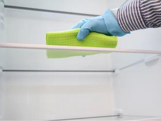 Mano enguantada que utiliza un paño verde brillante para limpiar un estante de vidrio del refrigerador.