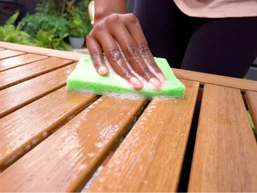 Persona limpiando una mesa de patio de madera con una esponja verde.