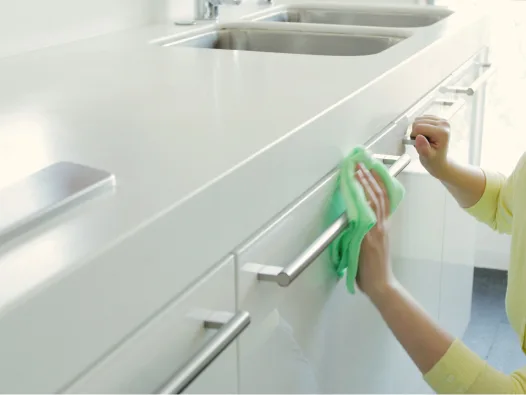 Hands wiping down stainless steel counter handles on white cabinets under sink with green cloth.