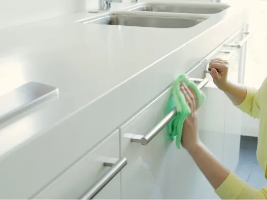 Manos limpiando las manijas de acero inoxidable de los gabinetes blancos debajo del fregadero con un paño verde.