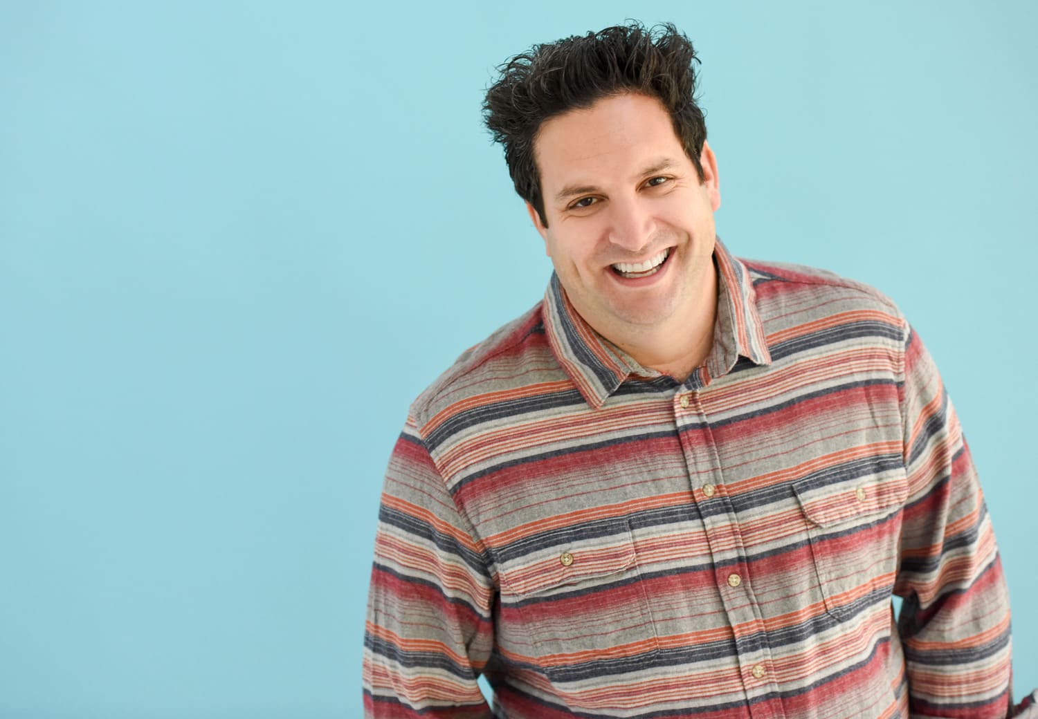 Man with tall dark hair smiling against a light blue background while wearing a striped button up shirt
