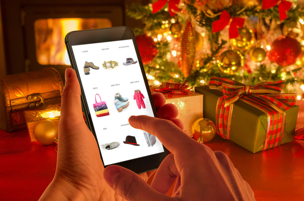 A person is browsing an online shop on a mobile phone with a christmas tree and presents in the background.