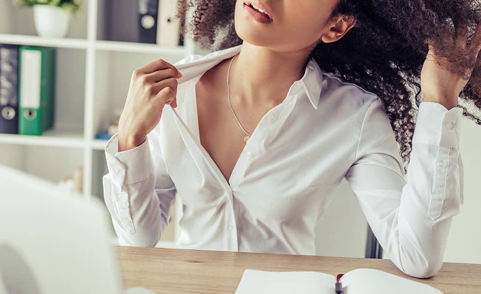 Woman at a desk pulling at her shirt collar while feeling warm.
