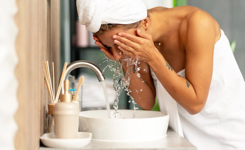 Woman wrapped in a white towel washing her face at a bathroom sink.