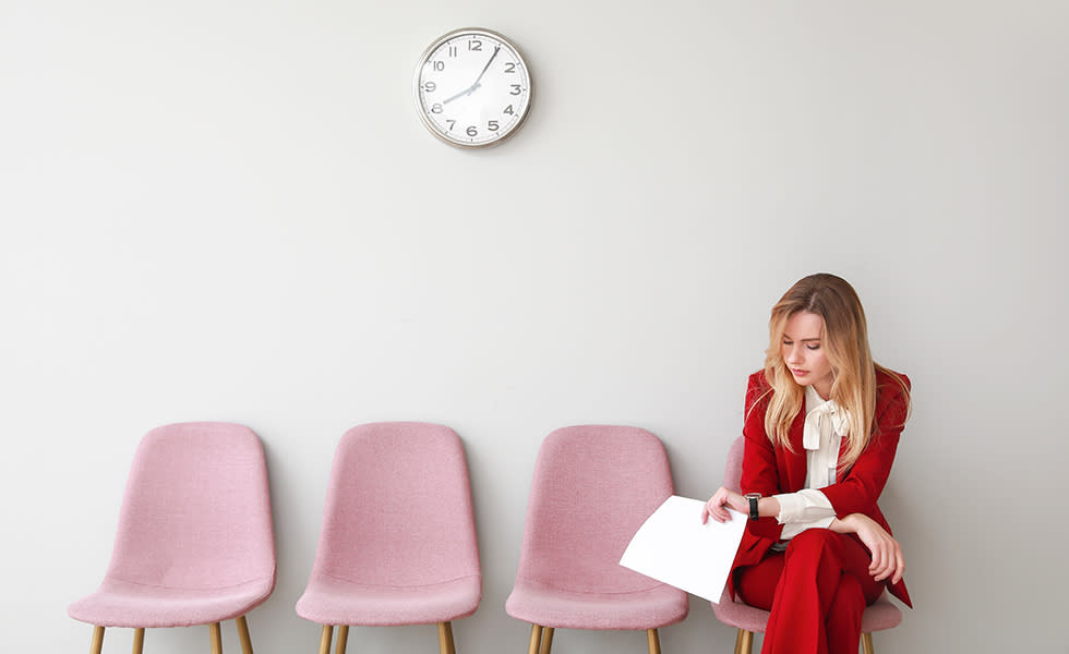 Woman in a red suit sitting in a waiting room holding papers beneath a wall clock.