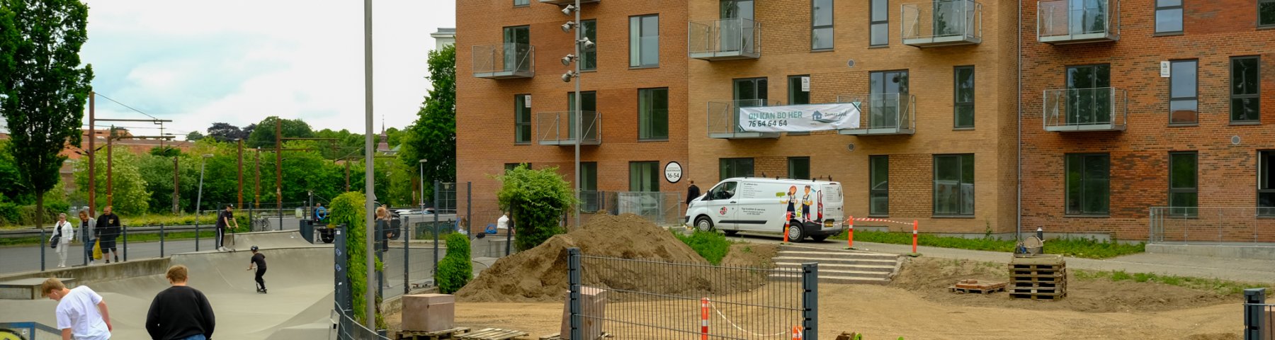 Skatere løber på en skaterbane til højre i billedet. Til venstre ses en åben plads foran et etagebyggeri i fire etager. Det er Klostergaarden.