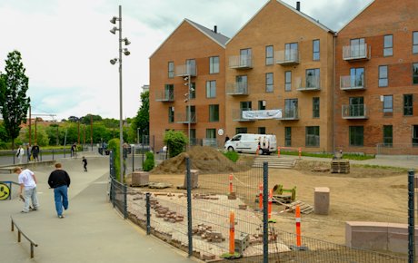 Skatere løber på en skaterbane til højre i billedet. Til venstre ses en åben plads foran et etagebyggeri i fire etager. Det er Klostergaarden.