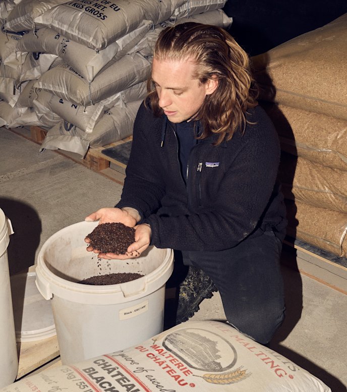 Philip Hulgaard sidder på hug i sit bryggeri foran en hvid spand og har løftet en stor håndfuld maltbyg op i sine hænder