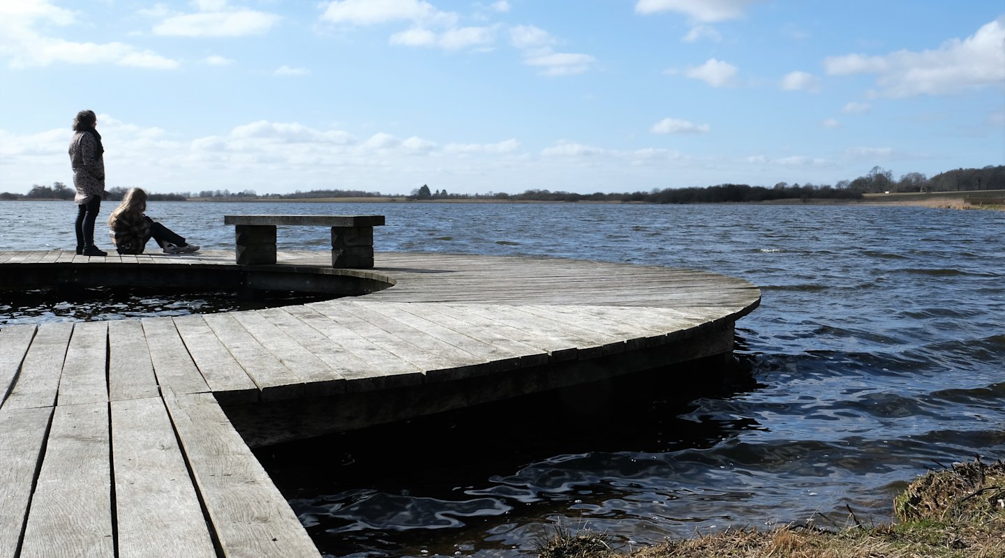 Ødis Sø fotograferet med en cirkelformet bro i forgrunden og til venstre i billedet. Bagest og til højre ses søen og i baggrunden landskabet, der omkranser søen. To personer opholder sig yderst på broen. Personen til venstre står op. Personen til højre sidder ned.