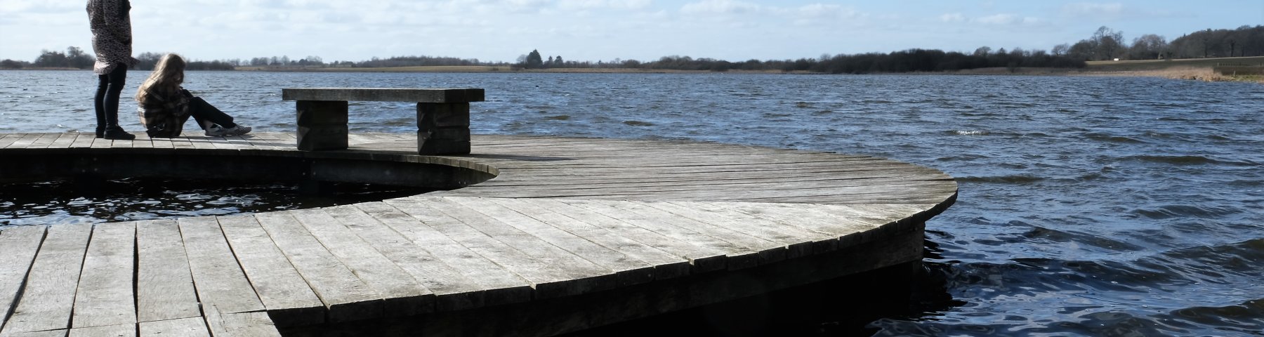 Ødis Sø fotograferet med en cirkelformet bro i forgrunden og til venstre i billedet. Bagest og til højre ses søen og i baggrunden landskabet, der omkranser søen. To personer opholder sig yderst på broen. Personen til venstre står op. Personen til højre sidder ned.