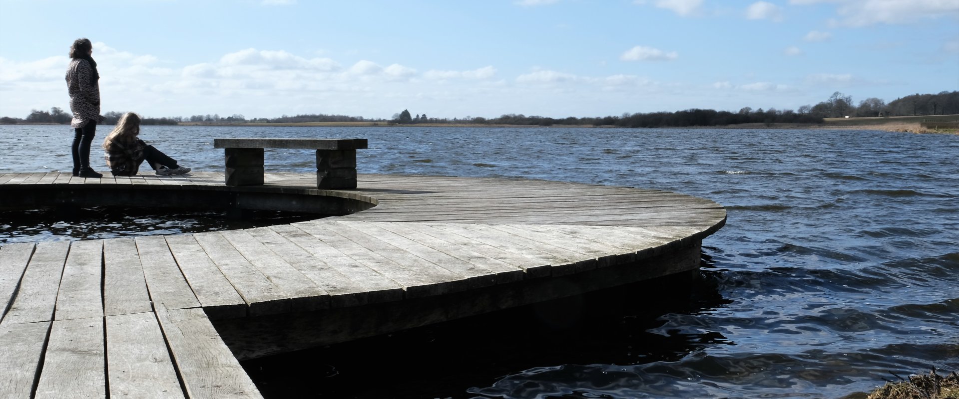 Ødis Sø fotograferet med en cirkelformet bro i forgrunden og til venstre i billedet. Bagest og til højre ses søen og i baggrunden landskabet, der omkranser søen. To personer opholder sig yderst på broen. Personen til venstre står op. Personen til højre sidder ned.