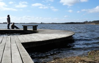 Ødis Sø fotograferet med en cirkelformet bro i forgrunden og til venstre i billedet. Bagest og til højre ses søen og i baggrunden landskabet, der omkranser søen. To personer opholder sig yderst på broen. Personen til venstre står op. Personen til højre sidder ned.