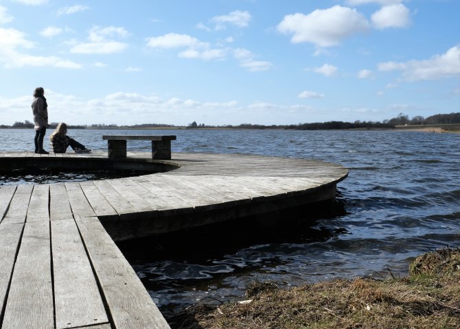 Ødis Sø fotograferet med en cirkelformet bro i forgrunden og til venstre i billedet. Bagest og til højre ses søen og i baggrunden landskabet, der omkranser søen. To personer opholder sig yderst på broen. Personen til venstre står op. Personen til højre sidder ned.