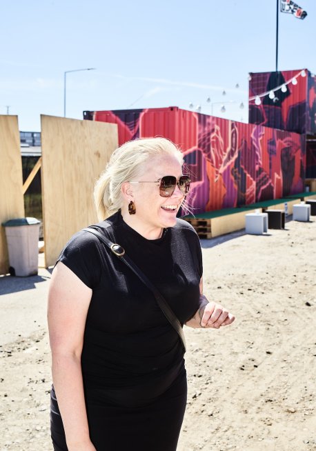 Rikke Molberg med solbriller på og stort smil og med graffitimalede containere ved udendørsscenen UJNFOR som baggrund