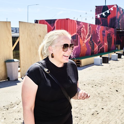 Rikke Molberg med solbriller på og stort smil og med graffitimalede containere ved udendørsscenen UJNFOR som baggrund