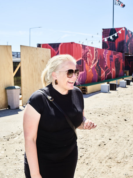 Rikke Molberg med solbriller på og stort smil og med graffitimalede containere ved udendørsscenen UJNFOR som baggrund