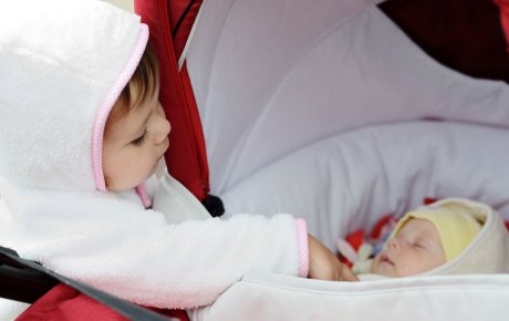 En baby ligger og sover i en barnevogn, så man kun kan se ansigtet. Et barn står og kigger på den sovende baby og rører forsigtigt ved babyens lille hånd