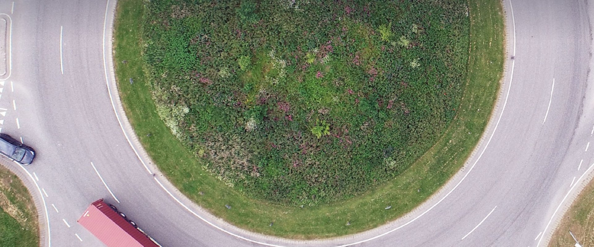 Dronefoto af en rundkørsel med en græsplæne i midten. En blå lastbil med rød anhænger er i rundkørslen nederst i billedet efterfulgt af en mørk personbil, der er på vej ind i rundkørslen i venstre side af billedet